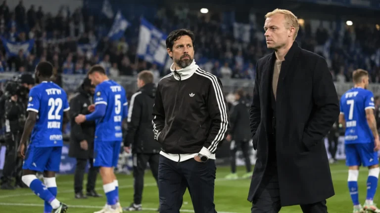 Medewerkers van Anderlecht staan langs de zijlijn terwijl spelers in blauwe shirts op de achtergrond lopen.