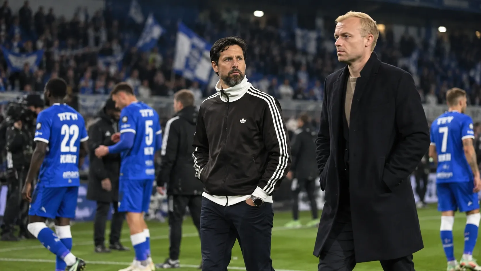 Medewerkers van Anderlecht staan langs de zijlijn terwijl spelers in blauwe shirts op de achtergrond lopen.