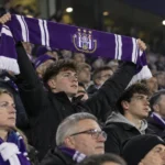 Een collectieve groep RSC Anderlecht-supporters op een avondtribune met één zichtbaar clubembleem op een sjaal.