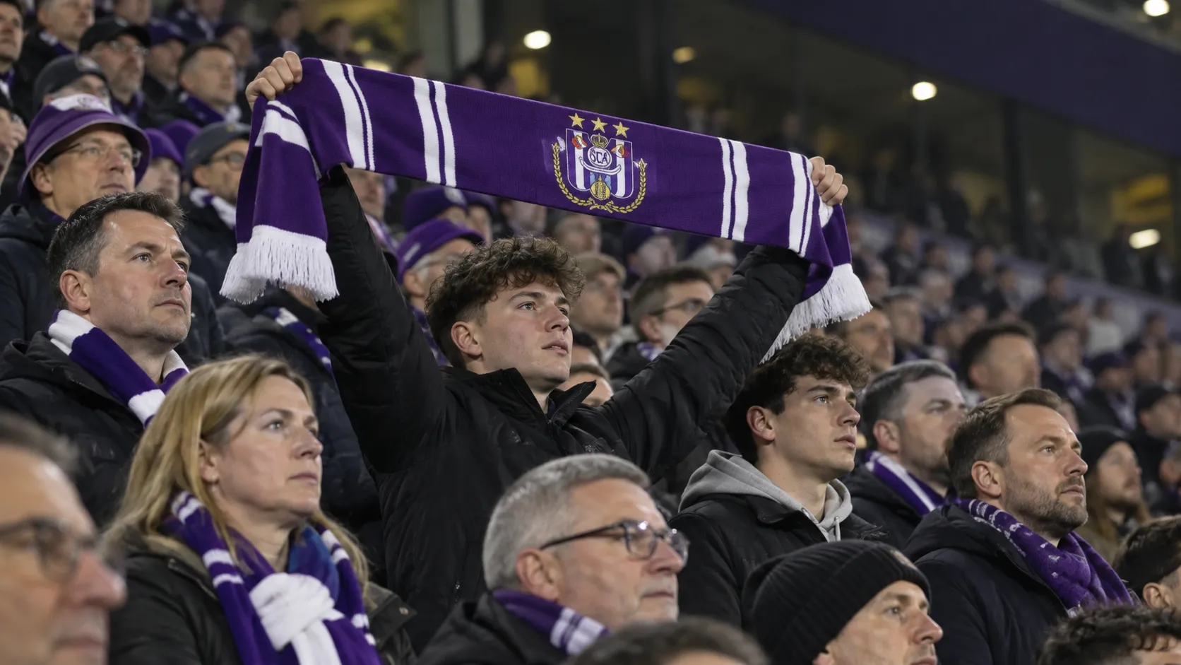 Een collectieve groep RSC Anderlecht-supporters op een avondtribune met één zichtbaar clubembleem op een sjaal.