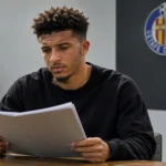 Jadon Sancho bekijkt een dossier in een neutrale vergaderruimte met een klein Getafe-embleem aan de wand.