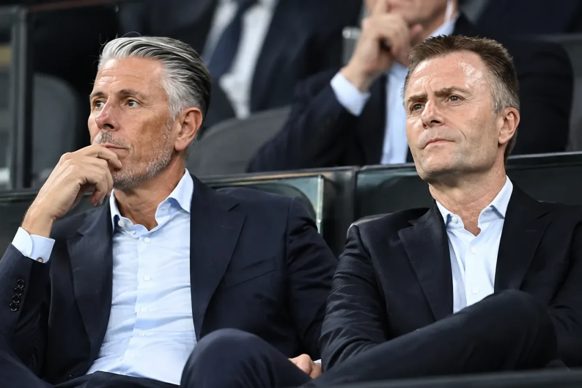 Twee clubbestuurders in donkere pakken op de tribune van een voetbalstadion.