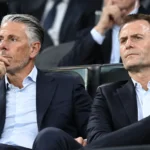 Twee clubbestuurders in donkere pakken op de tribune van een voetbalstadion.