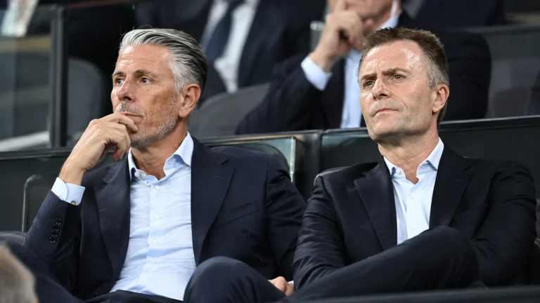 Twee clubbestuurders in donkere pakken op de tribune van een voetbalstadion.