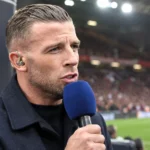 Een voetbalcommentator spreekt met een microfoon vanaf een perspositie in een stadion met een Manchester United-clubanker.