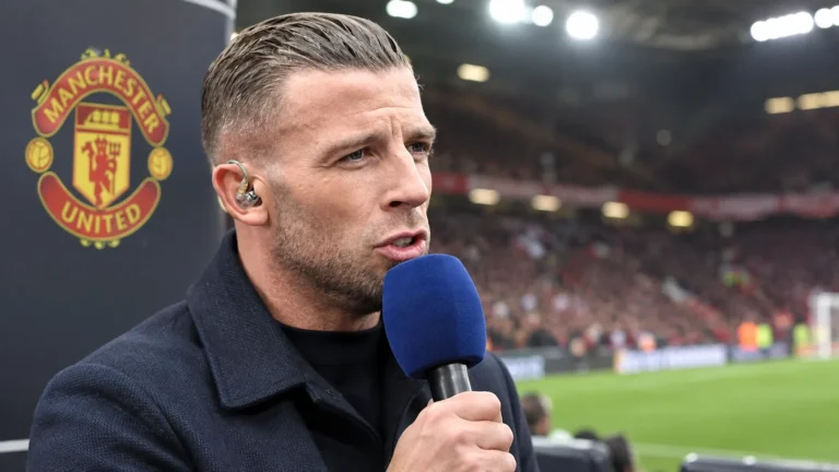 Een voetbalcommentator spreekt met een microfoon vanaf een perspositie in een stadion met een Manchester United-clubanker.