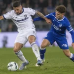 Twee voetballers in een blauw en een wit tenue tijdens een duel op het veld.