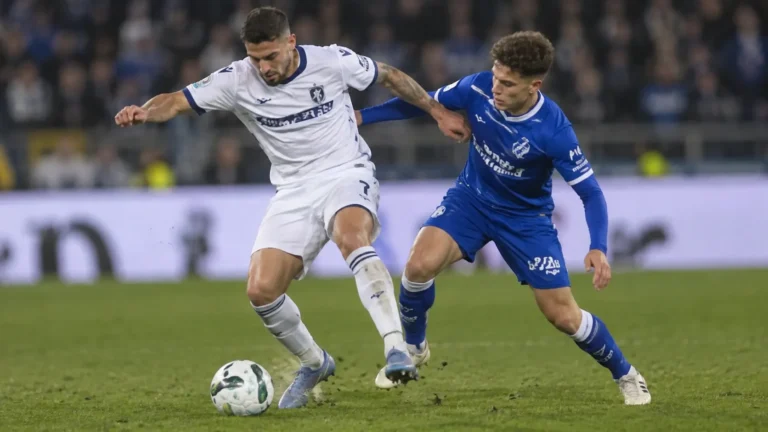Twee voetballers in een blauw en een wit tenue tijdens een duel op het veld.
