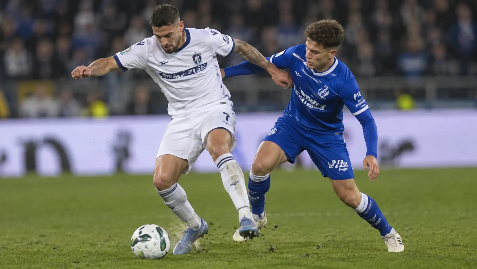 Twee voetballers in een blauw en een wit tenue tijdens een duel op het veld.