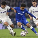 Een speler in blauw strijdt om de bal met twee spelers in witte wedstrijdtenues.