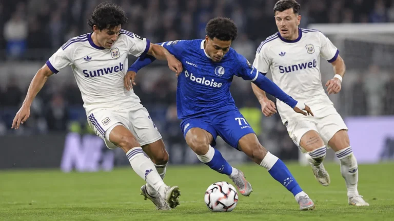 Een speler in blauw strijdt om de bal met twee spelers in witte wedstrijdtenues.