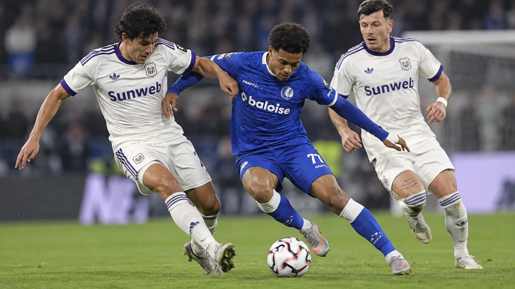 Een speler in blauw strijdt om de bal met twee spelers in witte wedstrijdtenues.