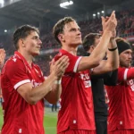 Een groep voetballers in rode shirts applaudisseert langs de rand van het veld.