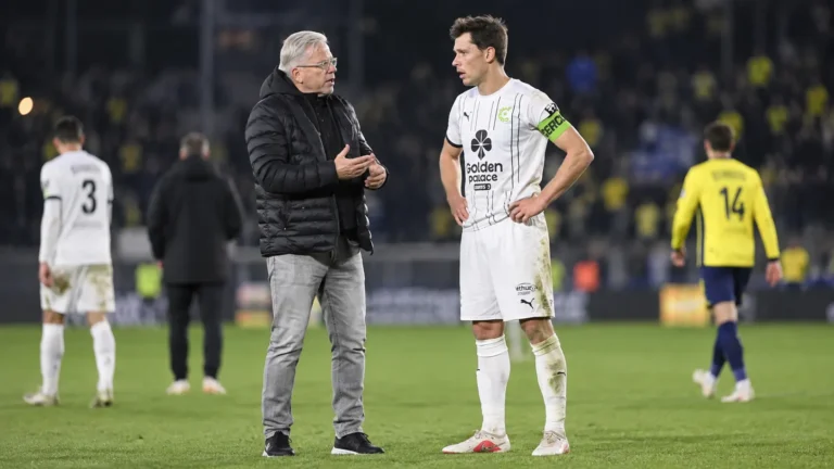 Een coach in zwarte jas spreekt na de wedstrijd op het veld met een speler van Cercle Brugge in wit tenue.
