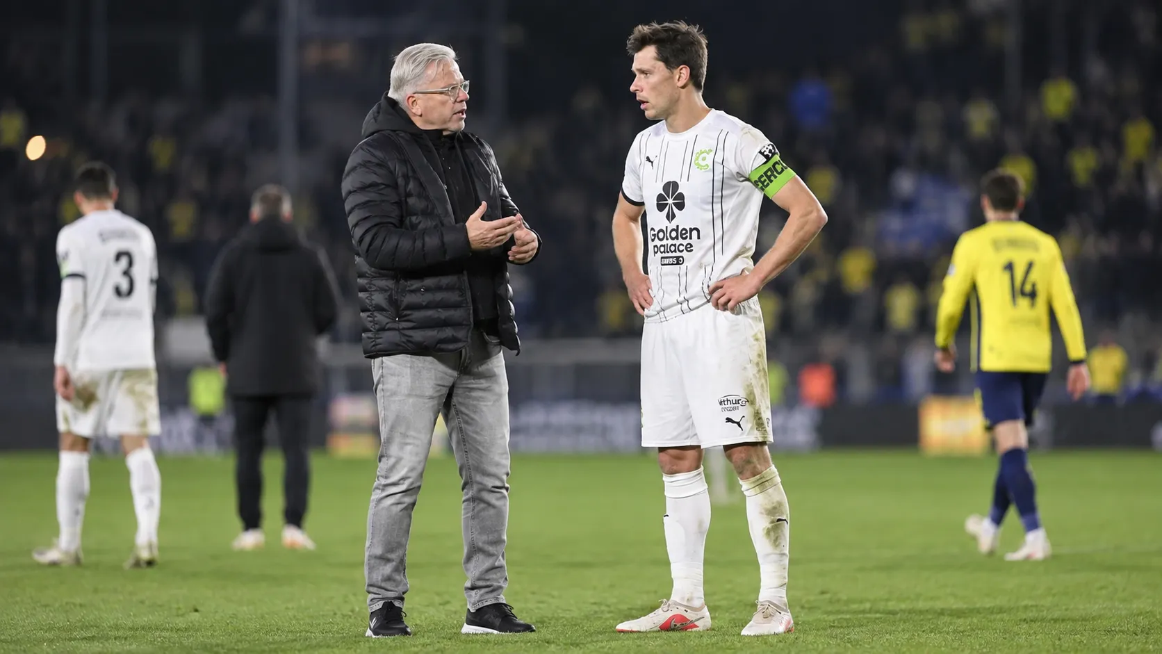 Een coach in zwarte jas spreekt na de wedstrijd op het veld met een speler van Cercle Brugge in wit tenue.