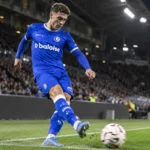Een voetballer in blauw tenue neemt bij avondlicht een hoekschop in een vol stadion.