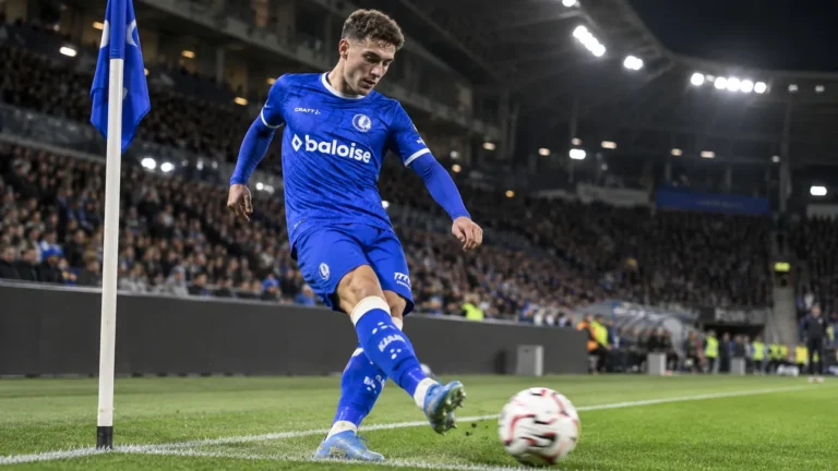 Een voetballer in blauw tenue neemt bij avondlicht een hoekschop in een vol stadion.