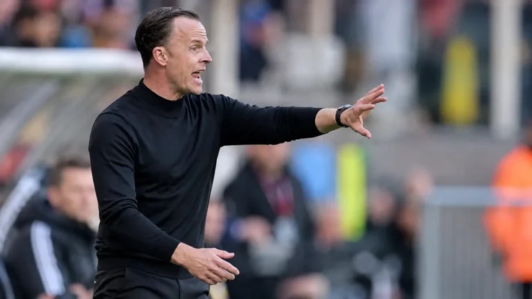 Een coach in een donkere trui geeft aanwijzingen langs de zijlijn.