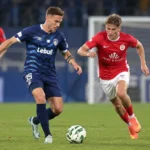Een speler in blauw strijdt om de bal met een tegenstander in rood op een voetbalveld.