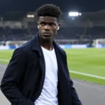 Aaron Bibout wandelt in een donkere jas langs de rand van een stadion.