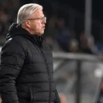 Lars Friis in een zwarte jas langs de zijlijn van een voetbalveld.