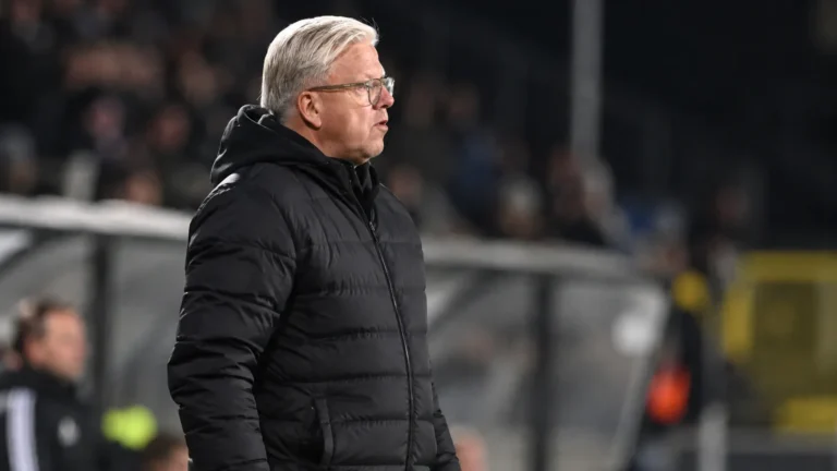 Lars Friis in een zwarte jas langs de zijlijn van een voetbalveld.