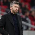 Michael Carrick staat in een donkere jas langs de zijlijn van een voetbalveld.