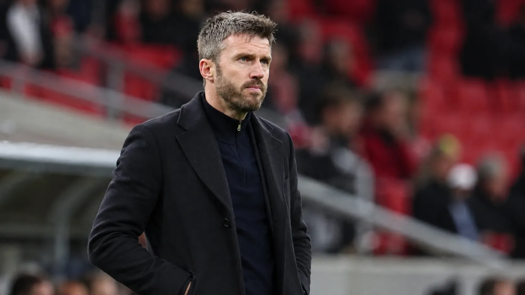 Michael Carrick staat in een donkere jas langs de zijlijn van een voetbalveld.