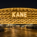 De goud verlichte Allianz Arena met de naam Kane op de gevel.