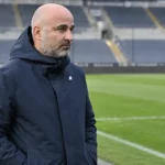 Een clubofficial in een donkere jas kijkt langs de rand van een leeg voetbalstadion.
