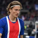 Een speler in een blauw-rood-wit wedstrijdshirt staat na een voetbalwedstrijd op het veld.