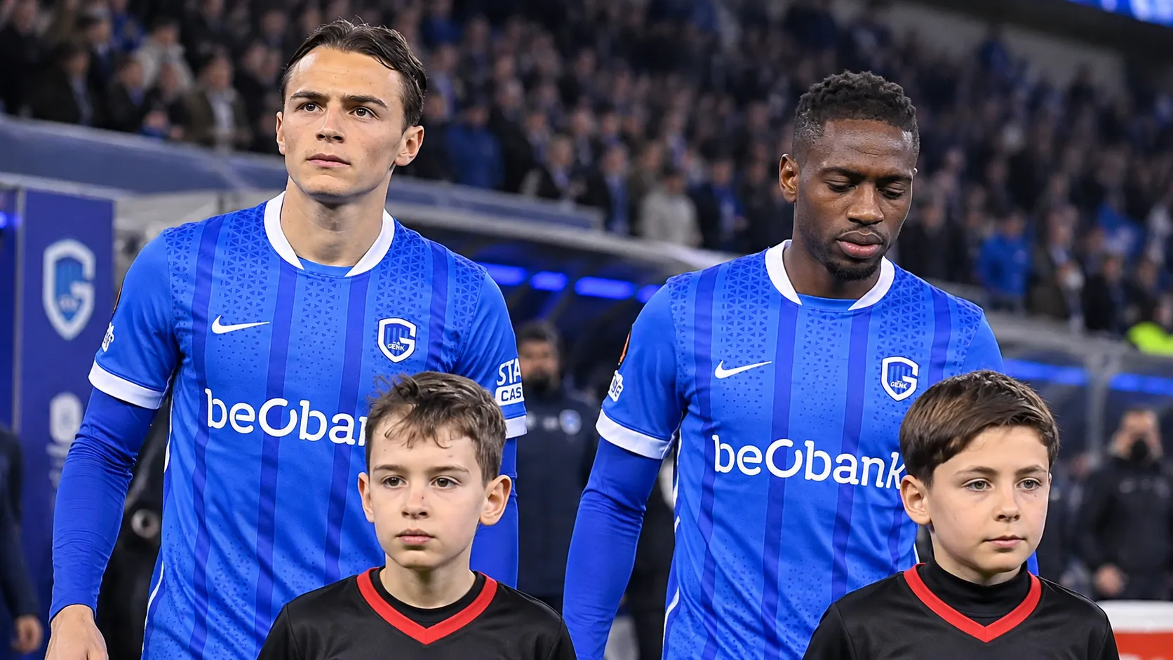Twee spelers van KRC Genk staan met jeugdmascottes op het veld voor een wedstrijd.