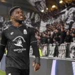 Cheick Keita in zwart voetbalshirt aan de rand van het veld voor een tribune.