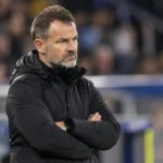 Een coach in een donkere jas observeert aandachtig het spel langs de zijlijn van een stadion.