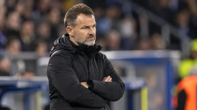 Een coach in een donkere jas observeert aandachtig het spel langs de zijlijn van een stadion.