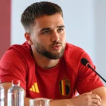 Nicolas Raskin in rood België-shirt aan tafel tijdens een mediamoment.