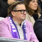 Een man met bril en paarse sjaal kijkt ernstig toe vanuit de tribune.