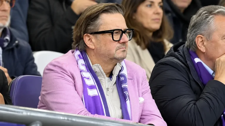Een man met bril en paarse sjaal kijkt ernstig toe vanuit de tribune.