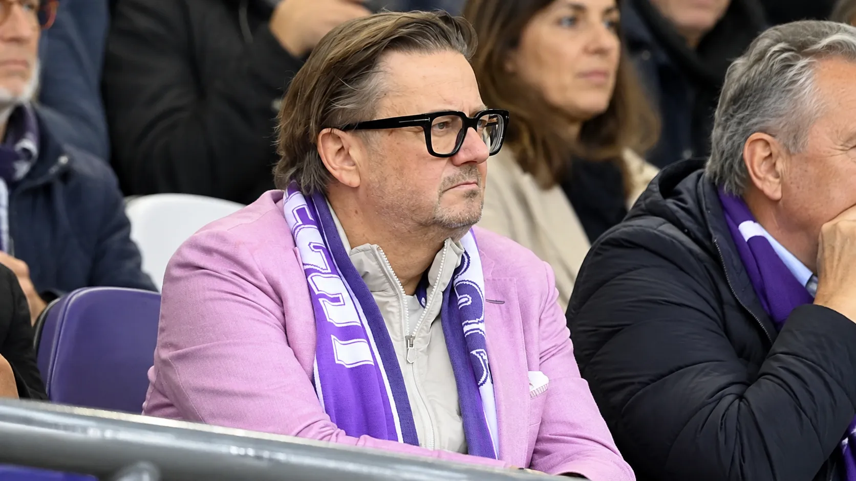 Een man met bril en paarse sjaal kijkt ernstig toe vanuit de tribune.
