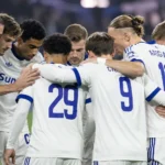 Een groep voetballers in witte tenues vormt een huddle op het veld.