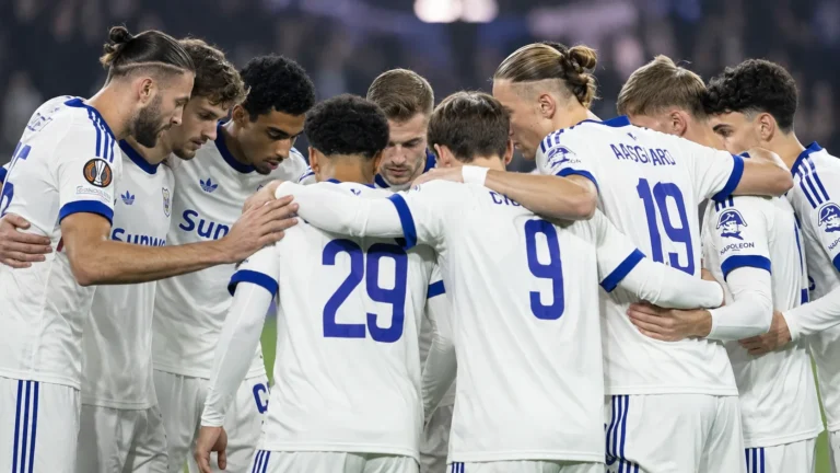 Een groep voetballers in witte tenues vormt een huddle op het veld.