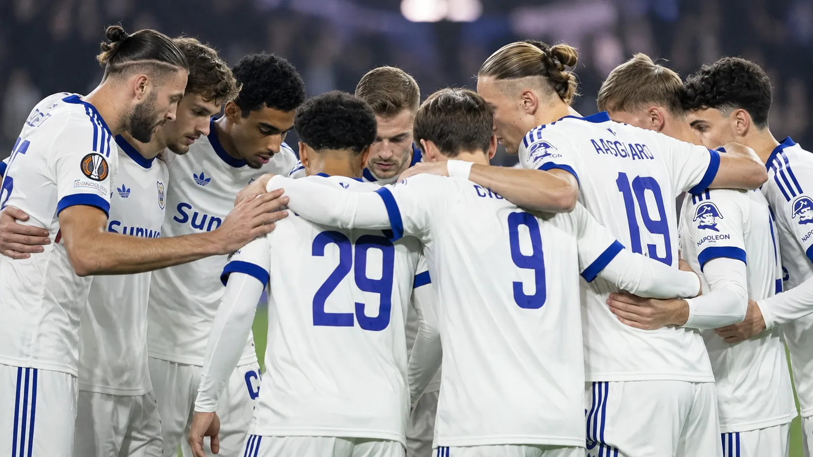Een groep voetballers in witte tenues vormt een huddle op het veld.