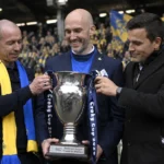 Drie clubvertegenwoordigers van Union Saint-Gilloise poseren met een beker in een stadion.