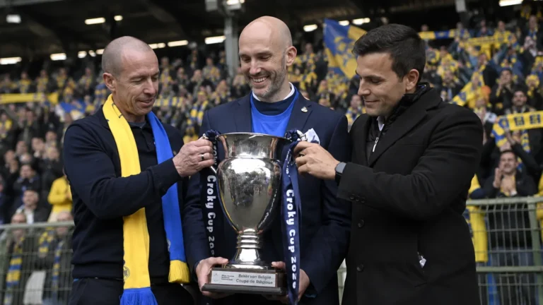 Drie clubvertegenwoordigers van Union Saint-Gilloise poseren met een beker in een stadion.