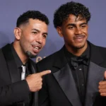 Twee mannen in formele avondkleding poseren voor een Ballon d’Or-achtergrond.