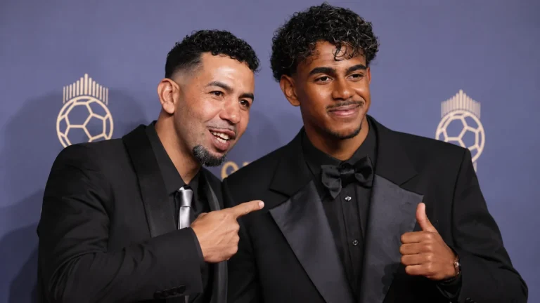 Twee mannen in formele avondkleding poseren voor een Ballon d’Or-achtergrond.
