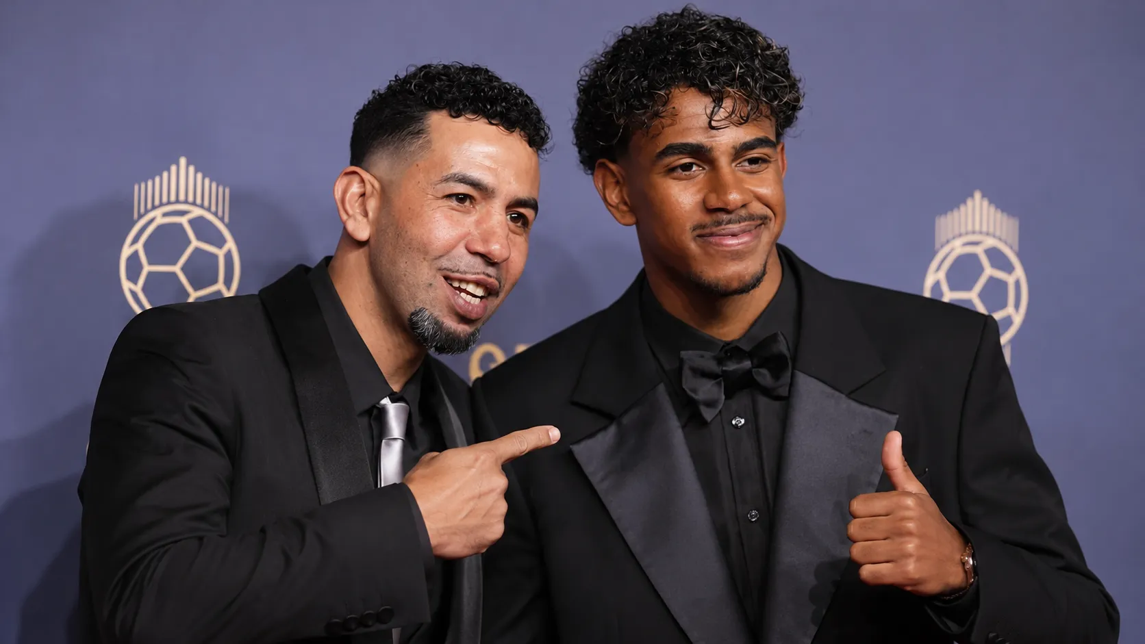 Twee mannen in formele avondkleding poseren voor een Ballon d’Or-achtergrond.