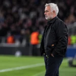 Een coach in donkere jas staat langs de zijlijn tijdens een voetbalwedstrijd.