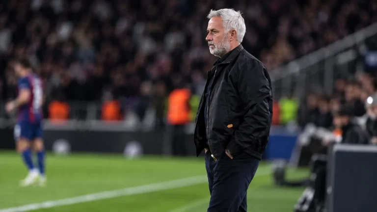 Een coach in donkere jas staat langs de zijlijn tijdens een voetbalwedstrijd.