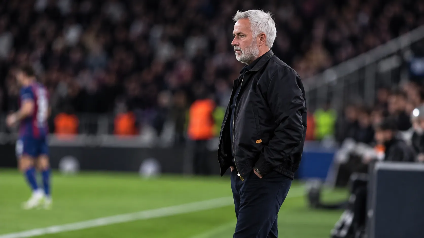 Een coach in donkere jas staat langs de zijlijn tijdens een voetbalwedstrijd.